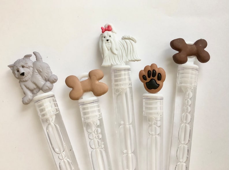 May include: Five clear plastic bubble wands with different dog-themed toppers. The toppers include a white dog with a pink bow, a brown bone, a gray dog, a brown bone, and a brown paw print.