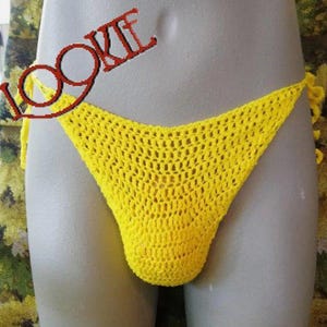 May include: A bright yellow crochet bikini bottom featuring an open-knit mesh pattern and side tie closures. The garment is displayed on a mannequin, and the word LOOKIE is printed in red text across the upper left portion of the item.