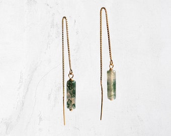 May include: A pair of gold-colored threader earrings. Each earring features a small, rectangular, green and white stone pendant suspended from a delicate chain. The earrings are displayed against a white background.