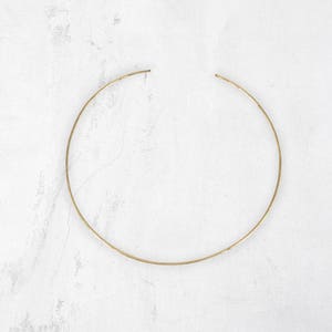 May include: A gold-colored choker necklace. The necklace is a simple, thin, circular band with open ends. The jewelry is set against a white, textured background.