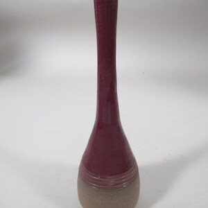 May include: A tall, slender vase with a narrow neck and a bulbous base. The upper portion features a glossy, deep burgundy glaze, while the lower section has a matte, textured, light brown finish separated by a series of horizontal ridges.