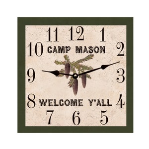 May include: A square, green-framed clock with a cream-colored face. The clock face features black Roman numerals, a black hour and minute hand, and a pine cone design with the text "Camp Mason" and "Welcome Y'all".