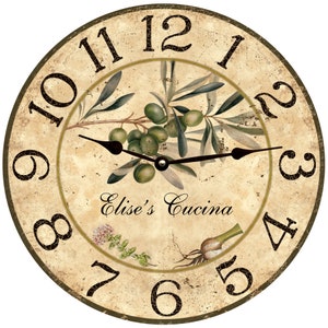 May include: A round, cream-colored clock with black Roman numeral hour markers and black hands. The clock face features a vintage-style illustration of olive branches with green olives, a sprig of thyme, and a garlic bulb. The text "Elise's Cucina" is printed in a cursive font.