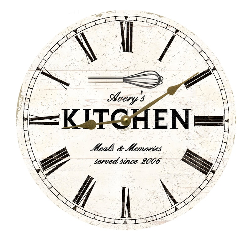 May include: White round kitchen clock with black Roman numeral markings and a gold-colored hand. The clock face has a distressed woodgrain texture and the words "Avery's Kitchen Meals & Memories served since 2006" are printed on the face.