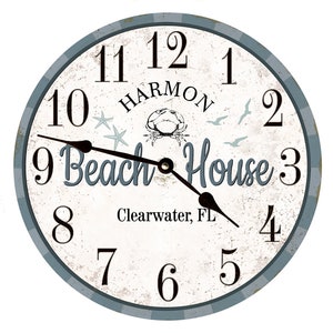May include: A white round clock with blue accents and black hands. The clock face has a distressed look and features the text "Harmon Beach House" with a crab and starfish design. The text "Clearwater, FL" is also included.