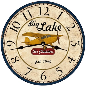 May include: A round wall clock with a cream-colored face, featuring black numerals and hands. The clock face displays a yellow airplane graphic with the text "Big Lake Air Charters Est. 1946". The clock has a dark blue border.