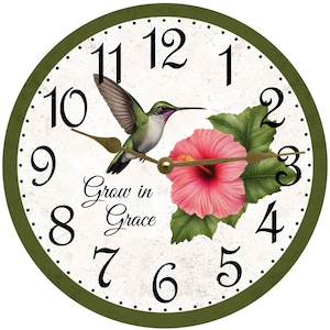 May include: Round wall clock with a green border, featuring a hummingbird and a pink hibiscus flower. The clock face has black numerals and hands, with the words "Grow in Grace" in script.