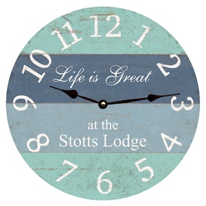 May include: Circular wall clock with a distressed finish. The clock face has a blue and teal striped pattern, white numerals, and black hands. The text reads "Life is Great at the Stotts Lodge."