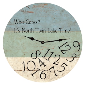 May include: A round wooden clock with a blue and white distressed wood background. The clock face has black numbers and hands. The clock face reads "Who Cares? It's North Twin Lake Time!"