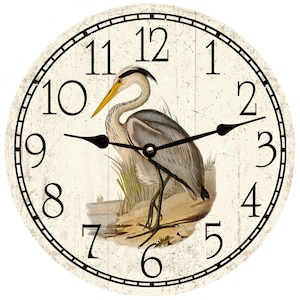 May include: A white clock face with black Roman numeral hour markers and black hands. The clock face features a detailed illustration of a grey heron standing on a branch with yellow reeds in the background.