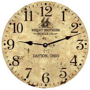 May include: A vintage-style clock face with Roman numerals and black hands. The clock face is cream-colored with a distressed look and features a black and white image of two people riding a bicycle. The text "WRIGHT BROTHERS BICYCLE CO." and "DAYTON, OHIO" is printed on the clock face.