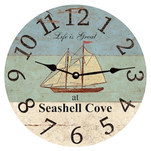 May include: A round wooden clock with a nautical theme. The clock face is painted in shades of blue and white with a sailboat in the centre. The numbers are black and the text reads "Life is Great at Seashell Cove".