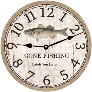May include: A white and brown wooden clock with black hands and Roman numerals. The clock face features a striped fish and the text "Gone Fishing Catch You Later..."