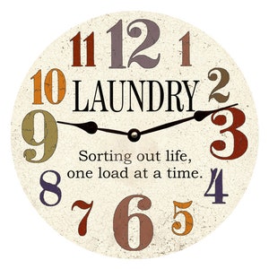 May include: A round, white clock with colourful numbers and the word "LAUNDRY" in black lettering. The clock face has a distressed, vintage look. The clock hands are black and point to the time. The clock has a quote that reads "Sorting out life, one load at a time."