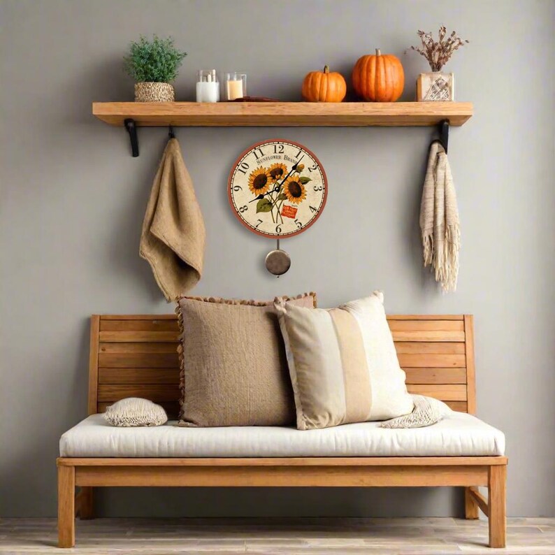 May include: A round wall clock with a sunflower design hangs on a gray wall. The clock has a pendulum and is surrounded by a wooden shelf with decorative items, including pumpkins, candles, and a plant. A wooden bench with pillows is below.