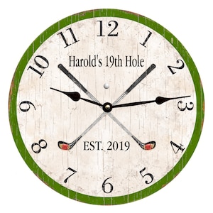 May include: A white and green round clock with black Roman numeral hour markers and black hands. The clock face has a distressed woodgrain texture and features a golf club design with the text "Harold's 19th Hole" and "EST. 2019".