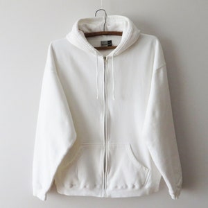 Könnte beinhalten: Ein weißer Kapuzenpullover mit Reißverschluss, Kordelzugkapuze und Vordertaschen. Der Sweatshirt hängt an einem Holzbügel vor einem schlichten weißen Hintergrund. Der Hoodie hat eine einfarbige, neutrale Farbe.