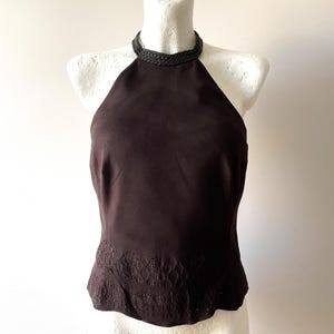 May include: A dark brown sleeveless halter top featuring a braided neckline detail and a decorative embroidered pattern along the bottom hem. The garment is displayed on a white mannequin, showcasing a fitted silhouette and a solid dark brown fabric.