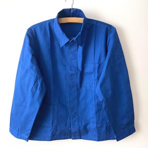 May include: A solid royal blue work jacket featuring a classic fold-down collar, long sleeves, and a button-front closure. The garment includes a single chest pocket on the left side and two lower front pockets, all constructed from a durable fabric.