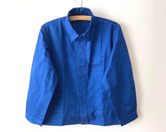 May include: A solid royal blue work jacket featuring a classic fold-down collar, long sleeves, and a button-front closure. The garment includes a single chest pocket on the left side and two lower front pockets, all constructed from a durable fabric.