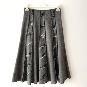 May include: A gray midi skirt with a flared silhouette and black and white embroidery. The skirt has a pleated design and a subtle sheen.