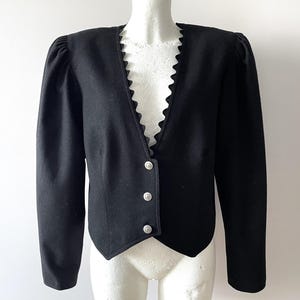 Könnte beinhalten: Schwarzer Blazer mit gewelltem Ausschnitt und drei silbernen Knöpfen. Die Jacke hat lange Ärmel mit gepolsterten Schultern, die eine strukturierte Silhouette schaffen. Diese Jacke im Vintage-Stil ist ein klassisches Oberbekleidungsstück.