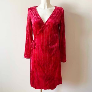 May include: A red velvet wrap dress with long sleeves and a V-neckline. The dress has a subtle shimmer and is adorned with delicate silver sequins.