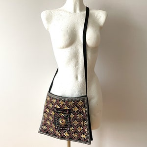 May include: Black crossbody bag with intricate embroidery. The bag features a square shape with a long, black strap. The embroidery includes gold, red, and white floral patterns, with a central square design.