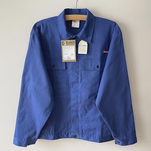 May include: A bright blue long-sleeve work jacket featuring a classic collar, a button-up front, and two chest pockets with button closures. The garment includes a brown rectangular product tag labelled PLANAM and a smaller white tag attached.
