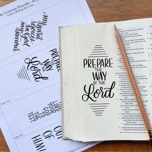 Könnte beinhalten: Ein weißes Blatt Papier mit schwarzen Kalligrafie-Designs aus dem Lukasevangelium, neben einem aufgeschlagenen Buch mit dem Text "PREPARE THE WAY OF THE LORD" auf der Seite und einem hellbraunen Holzstift, der diagonal auf dem Buch liegt.