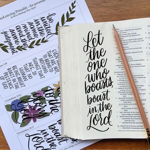 Może przedstawiać: Biała kartka papieru z czarnymi kaligraficznymi wersetami biblijnymi i ilustracjami kwiatowymi obok otwartej księgi z tekstem Let the one who boasts, boast in the Lord napisanym czarnym tuszem oraz jasnobrązowy ołówek leżący na stronie.