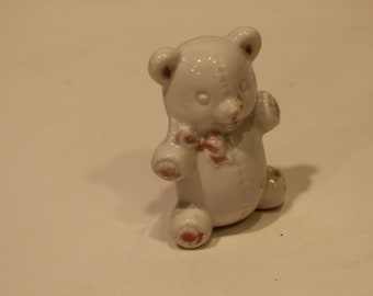 May include: A small white ceramic teddy bear figurine with a glossy finish. The bear features painted pink accents on its ears, nose, paws, and a small bow tied around its neck. It has molded stitch details on its chest and limbs, giving it the appearance of a plush toy.