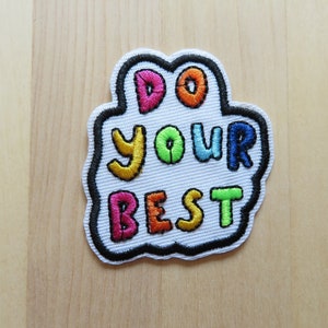 May include: Embroidered patch with the words "DO YOUR BEST" in colorful bubble letters. The patch has a white background with a black border and is set against a light wood grain surface.