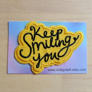 Puede incluir: Un parche bordado amarillo con las palabras "Keep Smiling You" en escritura negra. El parche tiene forma de nube y está sobre un fondo azul claro y morado.