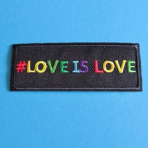 May include: A rectangular black patch with the embroidered text "#LOVE IS LOVE" in rainbow colors. The patch is set against a bright blue background, creating a vibrant contrast.