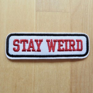 May include: A rectangular patch with the words "STAY WEIRD" embroidered in red on a white background. The patch has a black border with a white outline, set against a light wood grain surface.