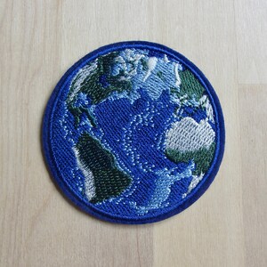 May include: Embroidered patch depicting Earth, featuring continents in green and white against a blue ocean. The patch is round with a dark blue border, showcasing a detailed representation of the planet.