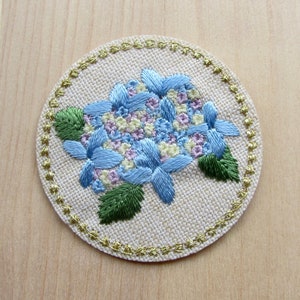 May include: Round embroidered coaster with a blue hydrangea design, green leaves, and yellow and purple accents. The floral design is on beige fabric, with a gold-coloured stitched border. The coaster is approximately 10 cm in diameter.