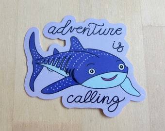 May include: A sticker featuring a cartoon whale shark in shades of blue and white, with the words "adventure is calling" in a whimsical font. The sticker has a light purple border.