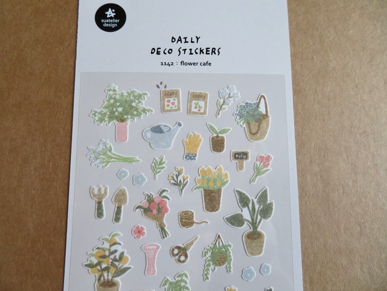 May include: A sheet of "Daily Deco Stickers" with a flower cafe theme. The stickers feature various gardening elements, including potted plants, flowers, gardening tools, and seed packets. The stickers are in soft pastel colors.