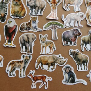 May include: A selection of die-cut stickers depicting African wildlife, such as a zebra, giraffe, lion, elephant, and rhinoceros. The stickers are in a watercolour style with detailed illustrations, arranged on a brown surface.