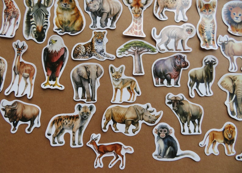 May include: A selection of die-cut stickers depicting African wildlife, such as a zebra, giraffe, lion, elephant, and rhinoceros. The stickers are in a watercolour style with detailed illustrations, arranged on a brown surface.