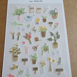 May include: A sheet of decorative stickers featuring various garden-themed illustrations. Includes potted plants, flowers, gardening tools, and labels with text like "tulip" and "basil". The color palette is soft, with pastel greens, pinks, and yellows.