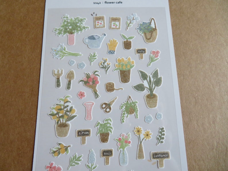 May include: A sheet of decorative stickers featuring various garden-themed illustrations. Includes potted plants, flowers, gardening tools, and labels with text like "tulip" and "basil". The color palette is soft, with pastel greens, pinks, and yellows.