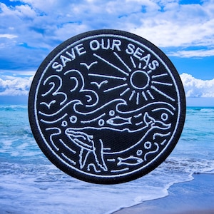 Puede incluir: Parche redondo negro con diseño bordado en blanco. El parche presenta una ballena, un sol, olas y el texto "SAVE OUR SEAS". El diseño incluye peces y pájaros. El parche está sobre un fondo de cielo azul y océano.