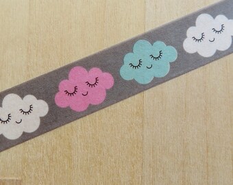 May include: Decorative washi tape featuring a repeating pattern of smiling cloud illustrations in pink, blue, and white on a gray background. The tape is angled across a light wood surface.