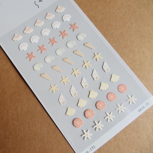 May include: A sheet of decorative stickers featuring various seashell and starfish designs. The stickers are in shades of peach, cream, and white, with some spiral and star patterns. The sheet has the text "DECO.21" and "NO.131".