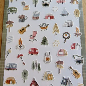 May include: A sheet of camping-themed stickers featuring illustrations of tents, trees, campfires, a guitar, a camera, and a camper. The stickers are in a variety of colors, including brown, green, red, and yellow. The stickers are on a white background.
