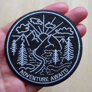 Puede incluir: Un parche redondo bordado en negro con un diseño de montaña y bosque en blanco. El parche presenta el texto "ADVENTURE AWAITS" en blanco, con un diseño de sol y nubes. El parche se sostiene en una mano.