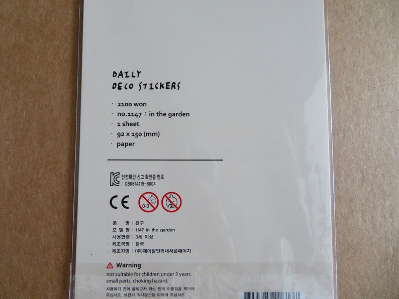 K&ouml;nnte beinhalten: Wei&szlig;e Verpackung von "DAILY DECO STICKERS" mit Text wie "no.1147: in the garden", Abmessungen von 92 x 150 mm und einem Warnhinweis zu Kleinteilen. Die Verpackung hat auch ein CE-Zeichen.